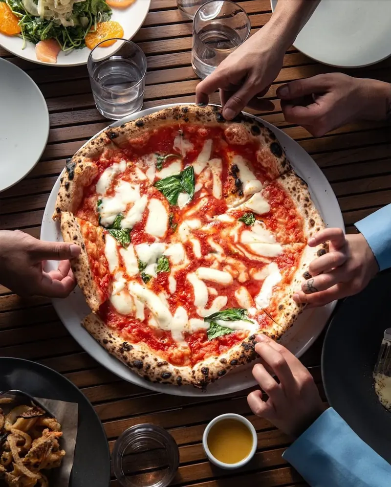 Overhead view of a Margherita with a blistered, leopard-spotted crust, shared by hands around a table